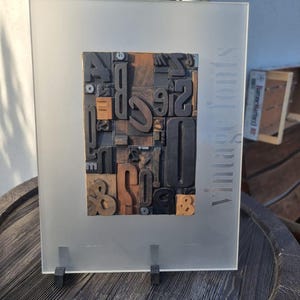 May include: A framed display of vintage wooden letterpress printing blocks. The blocks, in various fonts and sizes, are arranged in a rectangular pattern. The frosted glass frame has the words "vintage fonts" printed vertically. The display is supported by a black stand.