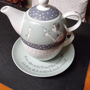 Può includere: Una teiera grigia e bianca con un piattino abbinato. La teiera ha un'illustrazione fantasiosa di una donna con un gatto e un uccello. Il piattino ha il testo "Prinz oder nicht Prinz? Das ist mir egal. Ich küss dich einfach noch einmal!" in tedesco.