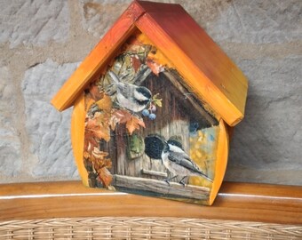 Op de afbeelding: Een houten vogelhuisje geverfd in tinten oranje en geel met een vogelhuisje scène met twee vogels op een tak. Het vogelhuisje staat op een rieten stoel.