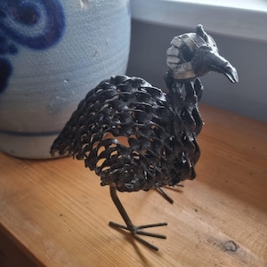 Op de afbeelding: Handgemaakte metalen sculptuur van een vogel met een getextureerd, opengewerkt lichaam en een gedetailleerde kop. De vogel is zwart met een zilverkleurig hoofddeksel en staat op dunne metalen poten. De sculptuur staat op een houten oppervlak.