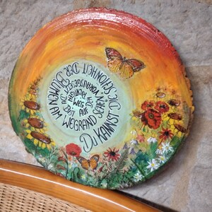 Puede incluir: Una rodaja de madera redonda con un diseño pintado de una mariposa, flores y el texto "Du kannst dich am Wegrand auf der großen Reise schon jetzt freuen". El fondo es un degradado de naranja, amarillo y verde.