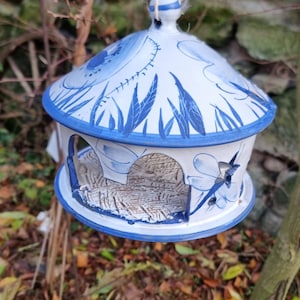 Op de afbeelding: Een blauwe en witte keramische vogelvoederhuisje met een koepelvormig dak en een decoratief ontwerp. De voederbak hangt aan een touwtje en heeft een kleine opening waar vogels toegang hebben tot het voedsel.
