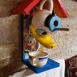 Pode incluir: Uma casa de pássaros de madeira pintada de vermelho, azul e branco, em forma de casa com telhado. A frente da casa é decorada com uma cabeça de pato de cerâmica com um olho azul e um bico amarelo. A cabeça de pato tem um buraco para os pássaros entrarem. Uma pequena xícara de chá azul e branca fica em uma prateleira abaixo da cabeça de pato.