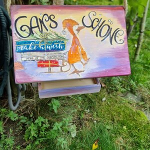 May include: Hand-painted wooden birdhouse with a whimsical design. The birdhouse features a drawing of a bird with a scarf and a sled, with the words "GANS SCHONE" and "make a wish" on the front. The birdhouse is suspended by a rope.