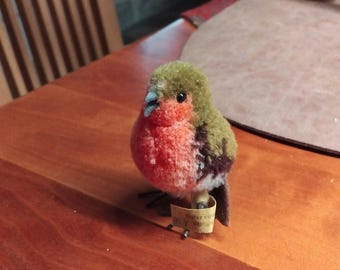 Könnte beinhalten: Eine kleine, flauschige Vogelfigur mit leuchtend roter Brust, olivgrünem Kopf und dunkelbraunen Flügeln. Der Vogel hat einen kleinen hellblauen Schnabel, schwarze Augen und dünne Drahtbeine. Ein kleines beiges Papieretikett ist an einem Bein befestigt.