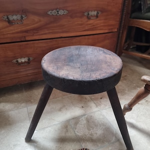 Könnte beinhalten: Ein kleiner dreibeiniger Holzhocker mit einer dicken, runden Sitzfläche in dunkler, verwitterter Optik und sichtbarer Holzmaserung. Der Hocker verfügt über drei einfache, konisch zulaufende Beine, die von der runden Sitzfläche ausgehen.