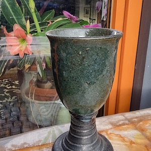 Könnte beinhalten: Ein dunkelgrüner Keramikkelch mit schwarzem Fuß. Der Kelch hat eine gesprenkelte Glasur und eine breite, runde Schale.