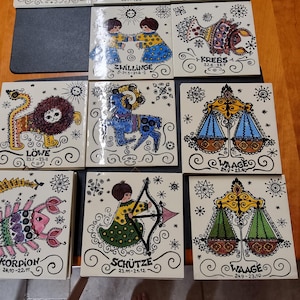 Op de afbeelding: Twaalf keramische tegels met kleurrijke illustraties van de sterrenbeelden. Elke tegel toont een ander sterrenbeeld met de bijbehorende data en naam in het Duits. De tegels zijn wit met zwarte contouren en hebben een verscheidenheid aan kleuren en patronen.