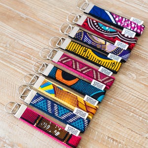 May include: A collection of eight fabric keychains featuring diverse geometric and abstract patterns in vibrant colours including blue, red, yellow, purple, and pink. Each keychain has a silver metal clasp and a small white tag labelled yamado.