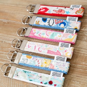May include: Six fabric keychains with metal rings and silver-toned hardware, each featuring a small white tag labelled "yamado". The keychains display various colourful patterns including lips, peace signs, eyes, rainbows, cats, and ladybugs.