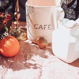 Op de afbeelding: Een stilleven met een witte keramische mok met de tekst "CAFE", een gouden zeeschelp en een feloranje vrucht. De mok heeft een gouden bloemenontwerp. Een witte, geometrische vaas en een gouden kandelaar zijn ook aanwezig.