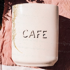 Puede incluir: Un recipiente de cerámica blanco con la palabra "CAFE" recortada en la parte delantera. El recipiente tiene forma rectangular y una superficie texturizada. Diseño botánico dorado en el lado izquierdo. Probablemente para almacenamiento en la cocina.