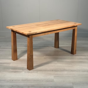 May include: A light brown wooden table with a rectangular top and four square legs. The table is made from solid wood, displaying the natural grain. The design is simple and functional. The table's dimensions are suitable for a dining room or kitchen. The table is approximately 150 cm long.