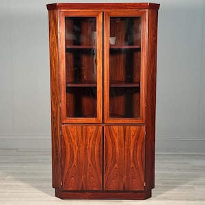 May include: A tall corner cabinet crafted from rich, dark-grained wood featuring a symmetrical design. The upper section includes two glass-panelled doors revealing interior shelving, while the lower section consists of two solid wood doors.