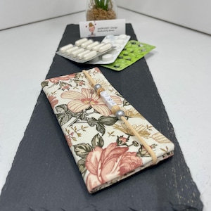 Könnte beinhalten: Ein rechteckiges Tablettenetui aus Stoff mit cremefarbenem Hintergrund und einem Blumenmuster aus rosa und orangefarbenen Blüten sowie grünen Blättern. Ein beiges Gummiband umschließt die Mitte, verziert mit Holzperlen und weißen Buchstabenperlen.