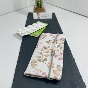 Könnte beinhalten: Ein rechteckiges Stoffetui mit einem hellen Muster aus braunen und rosafarbenen Blättern. Ein graues Gummiband umschließt das Etui, verziert mit kleinen Holzperlen und weißen Buchstabenperlen, die den Namen RAME in schwarzer Schrift bilden.