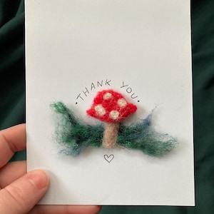 Puede incluir: Una tarjeta de felicitación hecha a mano con un diseño de seta de fieltro. La seta roja tiene manchas blancas y un tallo marrón, con follaje de fieltro verde y azul. Las palabras "gracias" están escritas encima de la seta, y un pequeño corazón está dibujado debajo.