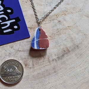 May include: A silver chain necklace with a pendant made from a piece of broken pottery. The pottery is red, blue, and white with a rough, uneven texture.