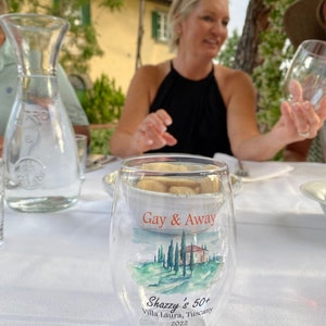 May include: A clear glass with the text "Gay & Away" and "Shazzy's 50+ Villa Laura, Tuscany 2022" printed on it. The glass features a watercolour image of a house and trees on a hillside.