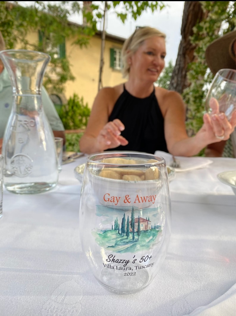 May include: A clear glass with the text "Gay & Away" and "Shazzy's 50+ Villa Laura, Tuscany 2022" printed on it. The glass features a watercolour image of a house and trees on a hillside.