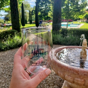 May include: A clear glass wine glass with a printed design of a Tuscan landscape and the text "Gay & Away Shazzy's 50+ Villa Laura, Tuscany 2022".