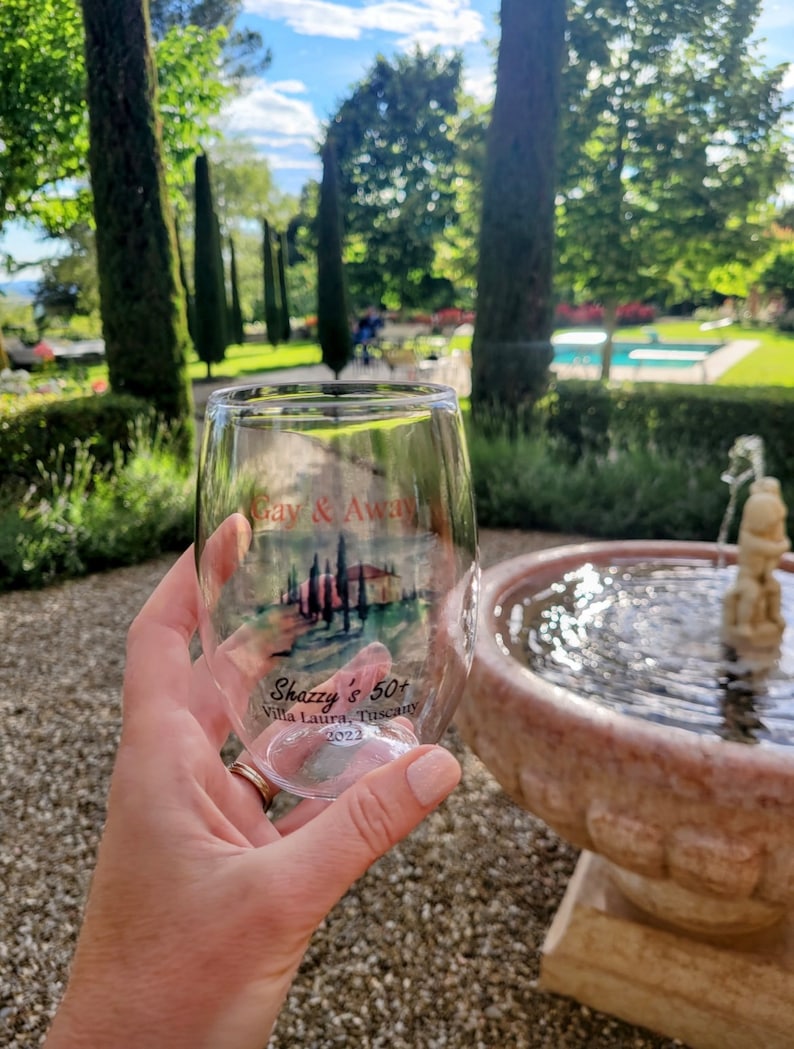 May include: A clear glass wine glass with a printed design of a Tuscan landscape and the text "Gay & Away Shazzy's 50+ Villa Laura, Tuscany 2022".
