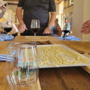May include: A clear glass with a printed design that says "Gay & Away" and "Shazzy's 50+ Villa Laura, Tuscany 2022". The glass is sitting on a wooden table with a plate of pasta in the background.