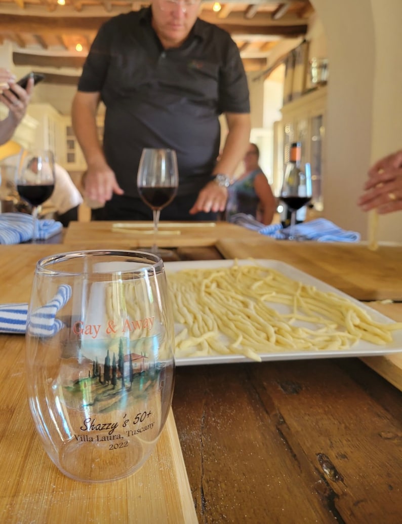 May include: A clear glass with a printed design that says "Gay & Away" and "Shazzy's 50+ Villa Laura, Tuscany 2022". The glass is sitting on a wooden table with a plate of pasta in the background.