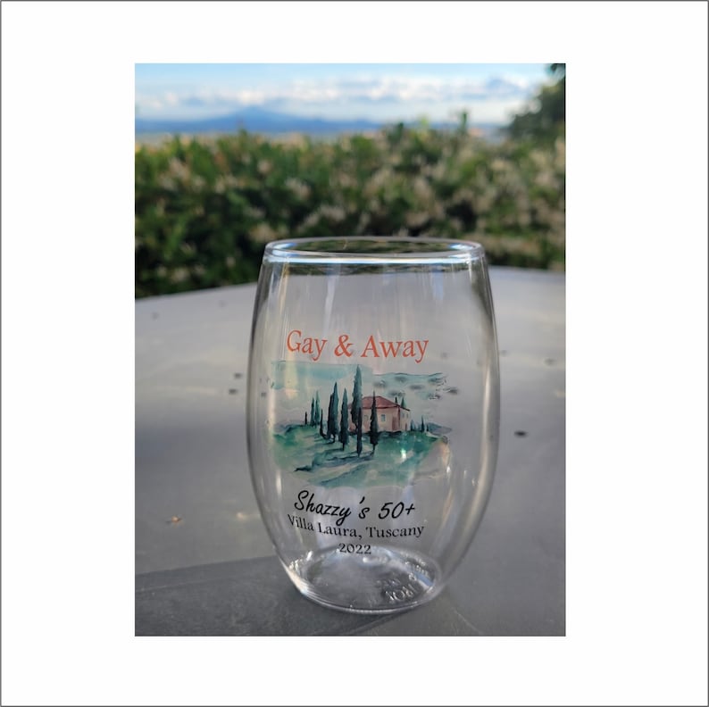 May include: A clear glass wine glass with a printed design of a Tuscan landscape with a house and trees. The text "Gay & Away" and "Shazzy's 50+ Villa Laura, Tuscany 2022" is printed on the glass.