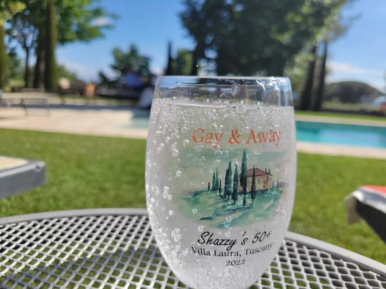 May include: A glass of sparkling water with bubbles in a clear glass with a painted design of a Tuscan landscape and the text "Gay & Away Shazzy's 50+ Villa Laura, Tuscany 2022".