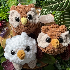 May include: Three crocheted owl plushies in brown, white, and brown with white accents. The owls have large black eyes and yellow beaks. The plushies are sitting on a wooden surface with green plants and crystals in the background.