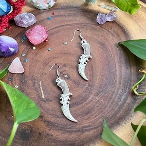May include: A pair of silver earrings shaped like daggers with intricate details. The earrings are displayed on a wooden surface with green leaves and crystals.