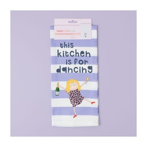 May include: A white kitchen towel with purple stripes and a cartoon illustration of a woman dancing with a glass of champagne. The text on the towel reads "This kitchen is for dancing."