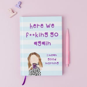 May include: A notebook with a light blue and white striped cover and the text "here we f**king go again" in purple. The notebook has an illustration of a woman holding a mug and the words "I mean good morning". A pink pen and two binder clips are also in the image.