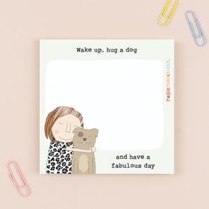 May include: A square notepad with the text "Wake up, hug a dog and have a fabulous day." The illustration features a person hugging a dog. The person is wearing a black and white patterned shirt. Pink, yellow, and blue paper clips are scattered around the notepad.