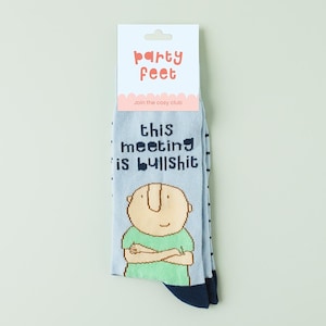 Op de afbeelding: Lichtblauwe sokken met de tekst "this meeting is bullshit" en een cartoonillustratie van een persoon met gekruiste armen. De sokken hebben een marineblauwe hiel en teen, en een wit label met de tekst "Party Feet".
