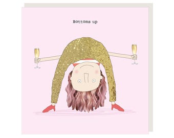 May include: A pink card with a cartoon illustration of a person in a gold sparkly outfit holding two glasses of champagne. The text "Bottoms up" is printed on the card.