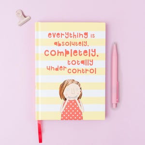 May include: A notebook with a yellow and white striped cover featuring the text "everything is absolutely, completely, totally under control" and an illustration of a person with brown hair wearing a red shirt with white stars, plus a pink pen.