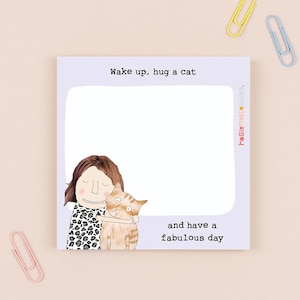 May include: A lilac sticky note pad with the text "Wake up, hug a cat and have a fabulous day." The pad features an illustration of a person hugging an orange tabby cat. The pad has a blank white space for notes.