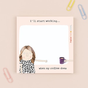May include: A square notepad featuring a cartoon illustration of a woman in a leopard print dress. The text reads "I'll start working... when my coffee does." A purple mug is held by a hand. Paper clips in pink, yellow, and blue are scattered around.