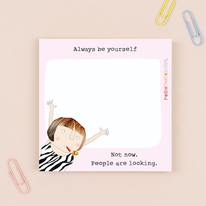 Op de afbeelding: Een roze notitieblok met de tekst "Always be yourself" en "Not now. People are looking." toont een cartoonillustratie van een persoon in een zebra-gestreept topje. Inclusief roze, gele en blauwe paperclips.