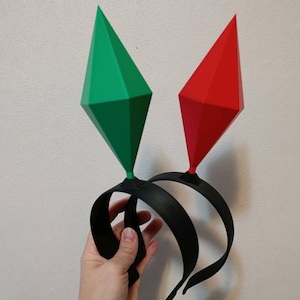 Puede incluir: Una diadema negra con dos formas geométricas, una verde y otra roja. Las formas de diamante están en posición vertical. La diadema es sostenida por una mano sobre un fondo blanco.