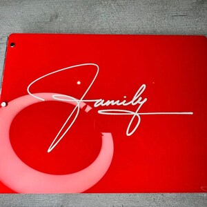 May include: A red, rectangular, acrylic sign with rounded corners and silver metal rivets. The sign has a white circle with a white cursive signature that says "Family".