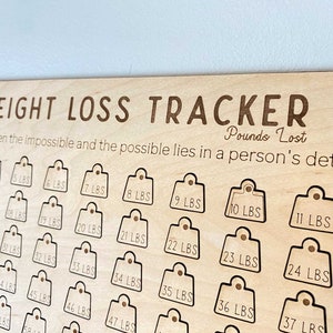 May include: Wooden weight loss tracker with a grid of 50 numbered tags, each representing a pound lost. The text "WEIGHT LOSS TRACKER" is at the top of the board, and the text "Pounds Lost" is below it. The text "The difference between the impossible and the possible lies in a person's determination" is below the "Pounds Lost" text.