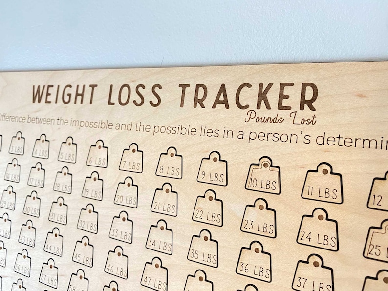 May include: Wooden weight loss tracker with a grid of 50 numbered tags, each representing a pound lost. The text "WEIGHT LOSS TRACKER" is at the top of the board, and the text "Pounds Lost" is below it. The text "The difference between the impossible and the possible lies in a person's determination" is below the "Pounds Lost" text.