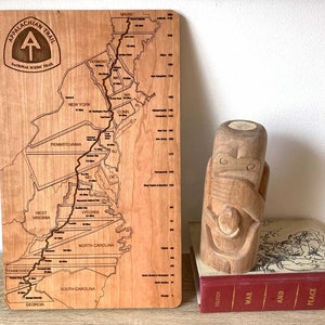 May include: A wooden map of the Appalachian Trail with state outlines and trail markings. The map is next to a carved wooden totem and a book titled "War and Peace". The map includes the text "Appalachian Trail National Scenic Trail".
