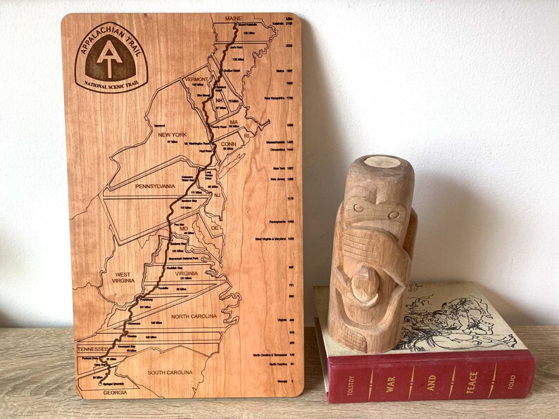 May include: A wooden map of the Appalachian Trail with state outlines and trail markings. The map is next to a carved wooden totem and a book titled "War and Peace". The map includes the text "Appalachian Trail National Scenic Trail".