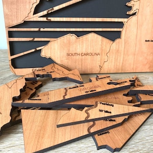 May include: A wooden map of the United States with individual state cutouts. The map is made of light brown wood with black backing. Several state pieces are separate, showing details like roads and distances in miles. The words "SOUTH CAROLINA" and "WEST VIRGINIA" are visible.