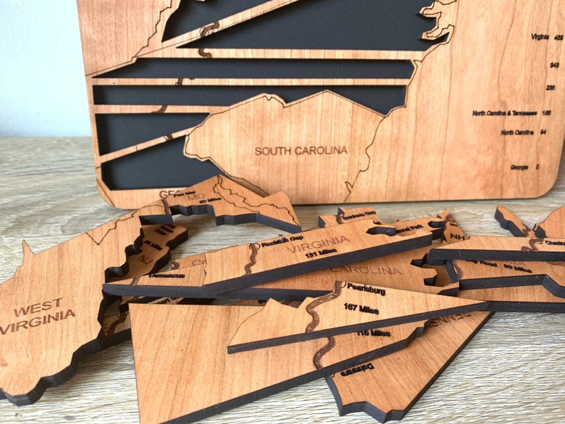 May include: A wooden map of the United States with individual state cutouts. The map is made of light brown wood with black backing. Several state pieces are separate, showing details like roads and distances in miles. The words "SOUTH CAROLINA" and "WEST VIRGINIA" are visible.