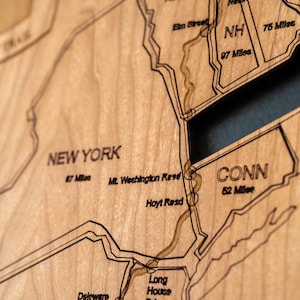 May include: Close-up of a wooden map of the northeastern United States, including New York, Connecticut, and Rhode Island. The map is etched with state lines, roads, and distances in miles. The wood has a light brown color.